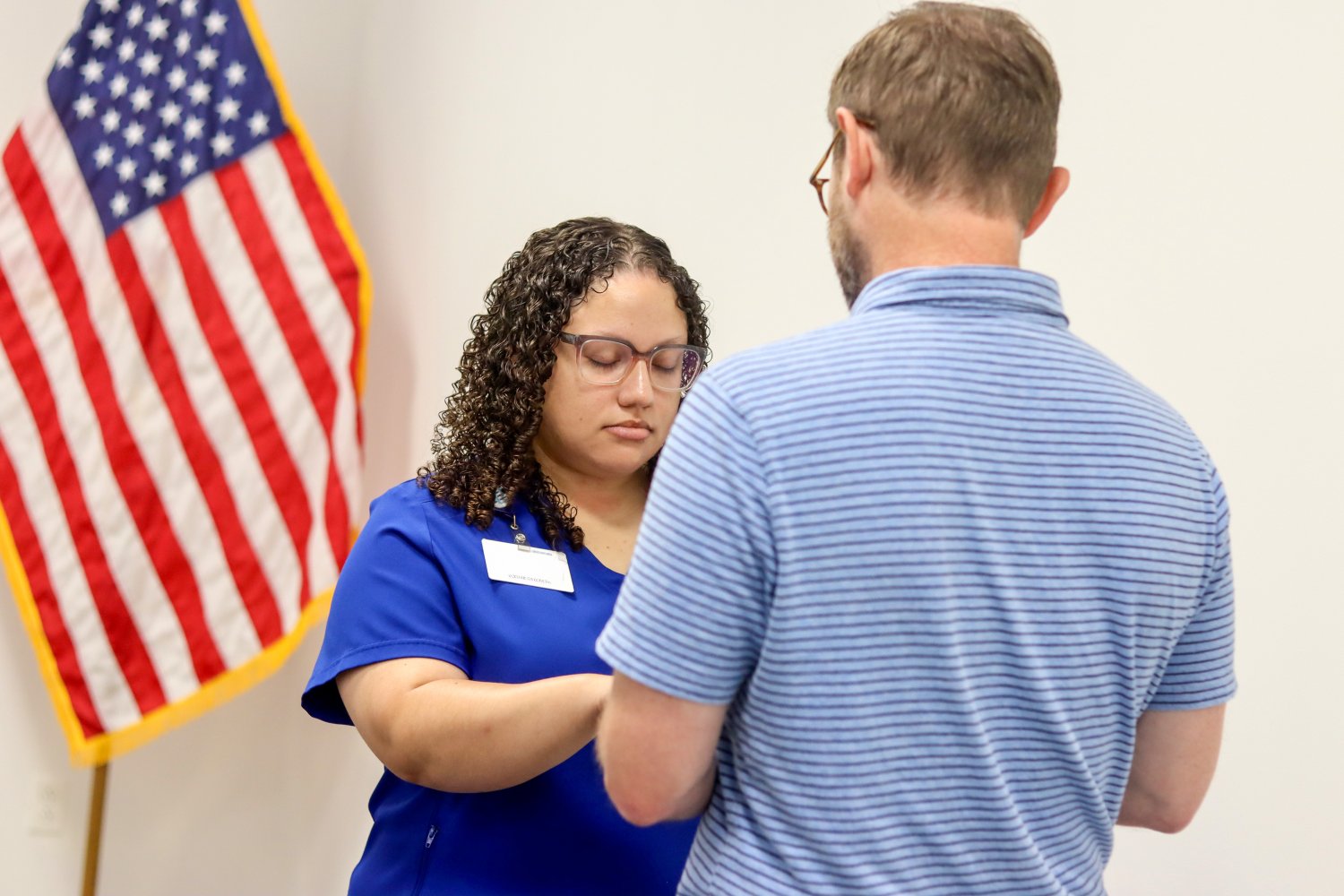 A female student with curly hair and glasses stands in front of the American flag as she holds hands with a speaker.