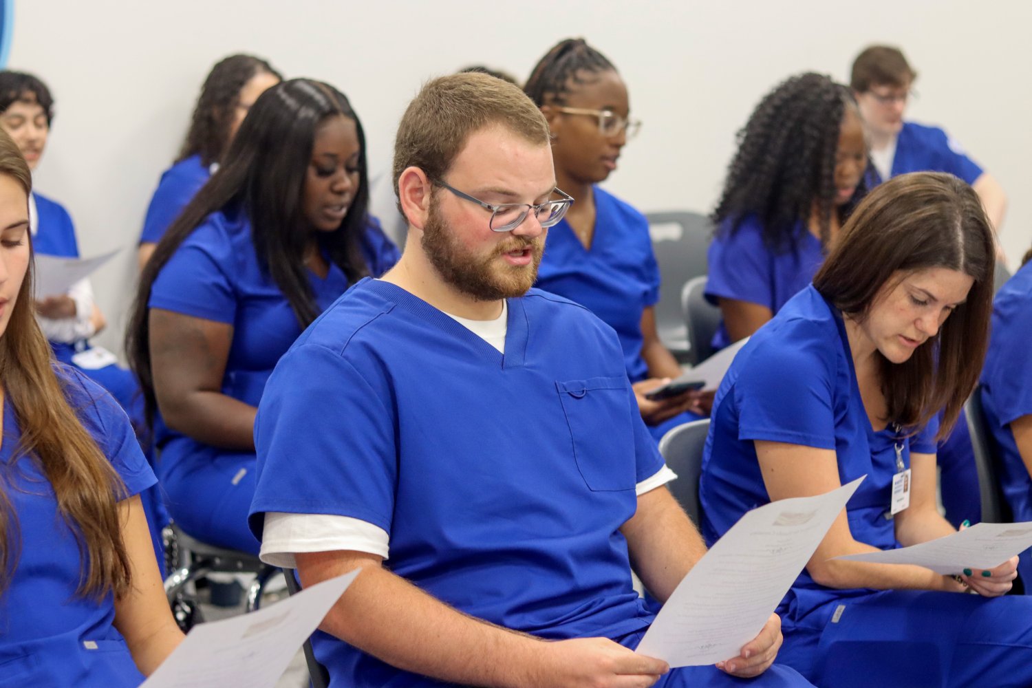 Students in blue scrubs read aloud from printed handouts during a formal event, seated in rows.