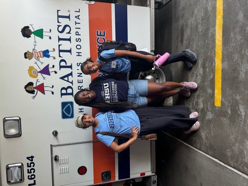 Three BUCOM students pose and smile in front of a Baptist Children’s Hospital ambulance. They wear BHSU HealthCORE t-shirts, backpacks, and casual attire, including a baseball cap and sneakers. The background features the ambulance’s colorful logo with cartoon children holding hands.