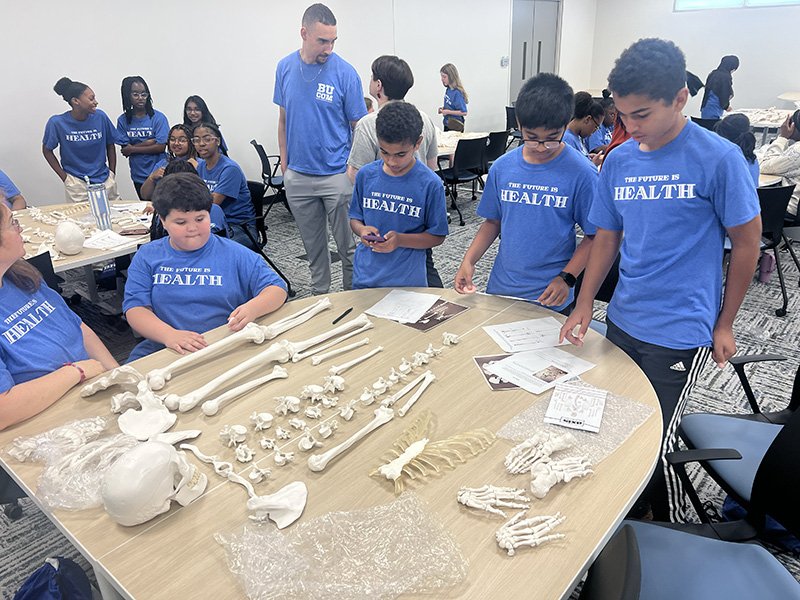  A group of students wearing matching blue shirts that read “The Future is Health” gather around a table displaying anatomical bone models. The table is covered with life-sized plastic bones including a skull, spine, pelvis, and limbs. Some students examine papers and booklets, while others closely observe the models. In the background, more students and adults are engaged in similar activities in a classroom setting.