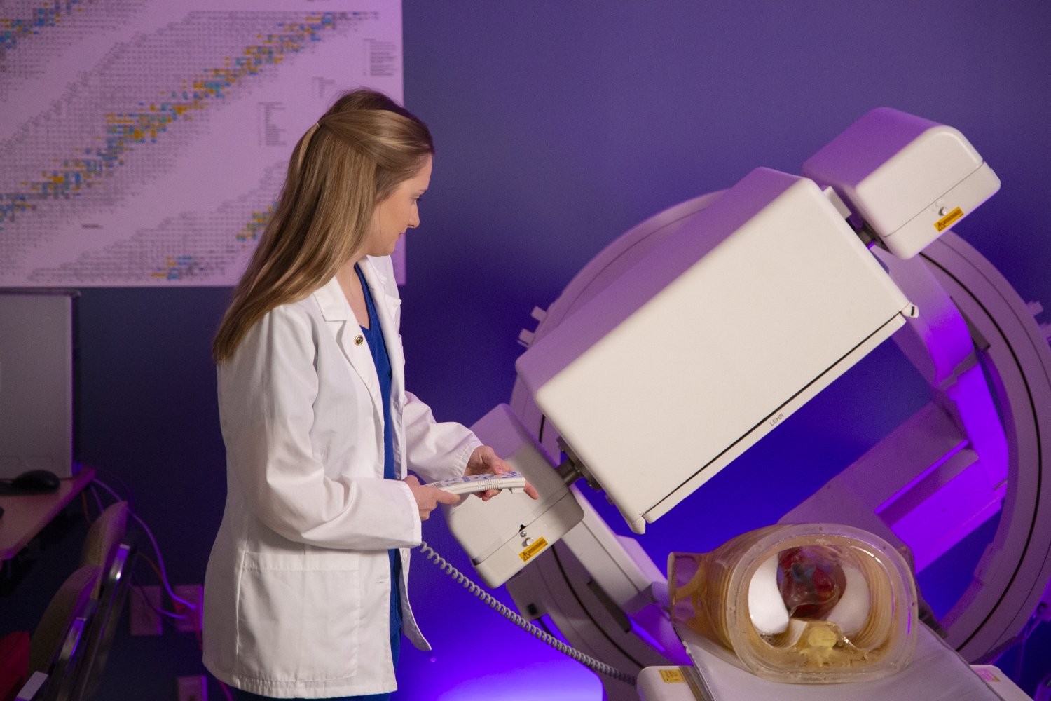 A female medical professional in a white lab coat operates a nuclear medicine imaging machine. A medical mannequin or anatomical model is positioned on the scanner table under the device. The background features a periodic table chart and purple ambient lighting.