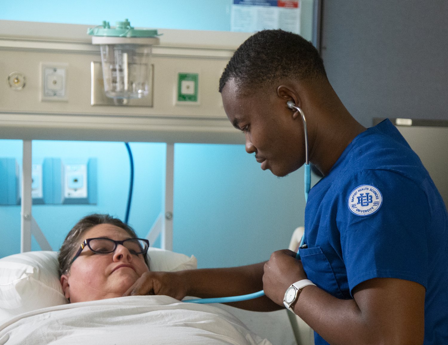 A nursing student in blue scrubs with a Baptist Health Sciences University patch listens to the chest of a patient lying in a hospital bed using a stethoscope. The patient is wearing glasses and appears calm. Medical equipment is visible on the wall behind them.