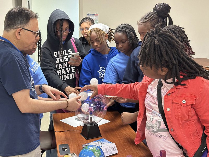 A group of students, mostly girls, gather around a table with a teacher demonstrating a plasma ball science experiment. The students are engaged, some touching the glass sphere to see the plasma filaments follow their fingers. The teacher is explaining, pointing at the device. The classroom setting includes papers, a small globe, and a smartphone on the table.