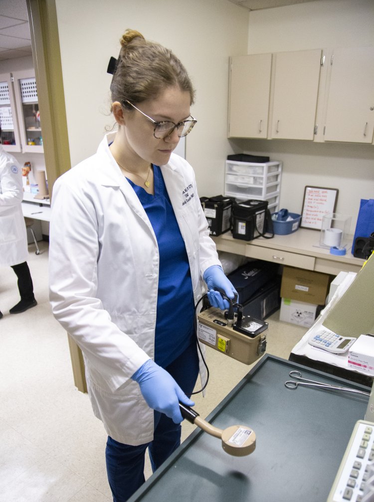 A female student in a lab coat and blue scrubs uses a handheld radiation detection device in a clinical lab setting. She wears safety gloves and glasses, and appears focused on scanning the surface of a table. Medical equipment and storage cabinets are visible in the background.