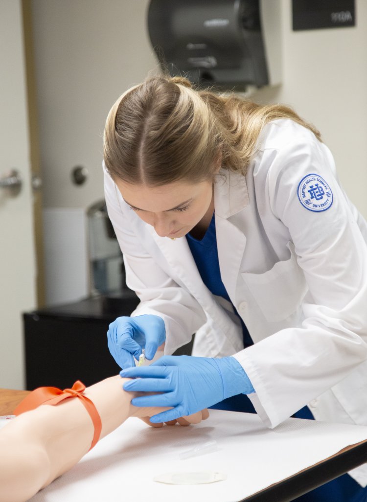 A female student wearing a white lab coat with the Baptist Health Sciences University logo and blue gloves practices an injection or IV placement on a medical training arm in a clinical skills lab. She is focused on the task, demonstrating hands-on healthcare training.