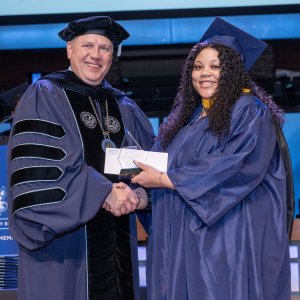 A smiling graduate in a navy blue cap and gown shakes hands with a university official in academic regalia while receiving an award on stage at a commencement ceremony.