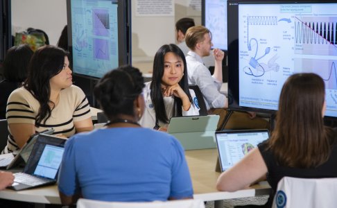 Medical students collaborate in a small group classroom setting, surrounded by large digital screens displaying anatomical and physiological diagrams. They are engaged in discussion, using laptops and tablets. A female student in a white coat, centered in the image, looks attentively toward her peers. The environment reflects an interactive and technology-enhanced medical learning experience.