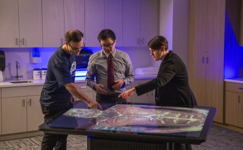 Three individuals—two men and one woman—interact with a high-tech digital anatomy table displaying a detailed human body. They are engaged in discussion and pointing to anatomical features, in a modern medical lab setting with cabinets, sinks, and soft blue lighting in the background. The scene highlights hands-on, tech-enhanced learning in a healthcare education environment. 