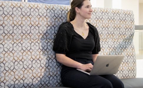 A woman sits on a patterned lounge bench with a laptop on her lap, looking to the side and smiling slightly. She is dressed in business-casual attire and appears to be working or studying in a modern, well-lit academic or office space. 