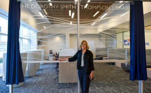 A woman stands in front of a glass wall bearing the inscription 'The Dorothy Smith Thompson Library.' The space behind the glass appears to be a modern, well-lit library or study area with comfortable seating and workstations. Blue curtains are drawn back on either side of the glass. The woman is wearing glasses and a dark outfit, smiling slightly at the camera.