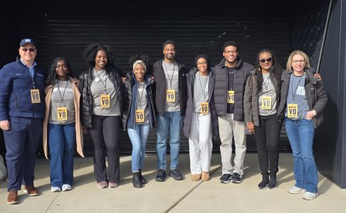 Group of ten adults standing side by side outdoors, smiling at the camera. Most wear gray BUCOM T-shirts layered with jackets and ‘King Day Volunteer’ badges on lanyards. They pose with arms around each other in front of a dark building entrance, suggesting a team volunteer event.