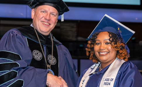 A graduate from Baptist Health Sciences University, dressed in a navy cap and gown with a white stole reading “Class of 2024,” smiles while shaking hands with a university official in academic regalia during the commencement ceremony.