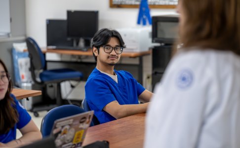 A student in blue medical scrubs sits at a classroom desk, looking toward a faculty member in a white coat speaking at the front of the room. Another student with a laptop is partially visible in the foreground, and computer equipment is in the background.