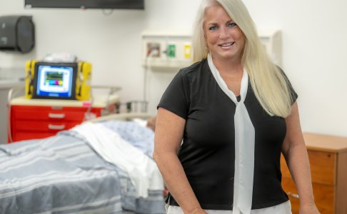  A smiling woman with long blonde hair stands in a medical simulation lab, wearing a black blouse with a white scarf-like detail. Behind her is a bed with a medical mannequin, a red emergency cart, and medical equipment.