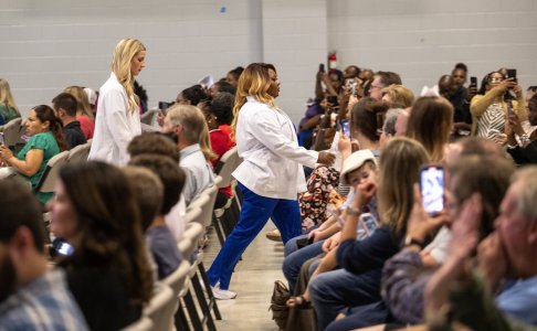 Two students in white coats walk down an aisle during a ceremony as family and friends seated on both sides take photos and cheer in support.