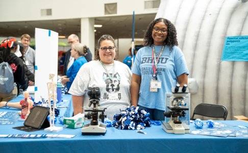 Two women stand behind a BHSU Healthcare booth, smiling at the camera. The table features anatomical models, microscopes, pom-poms, and brochures. One woman wears a BHSU Healthcare t-shirt, and the other wears a shirt with a stylized dog graphic. Behind them, a large inflatable colon exhibit is partially visible.