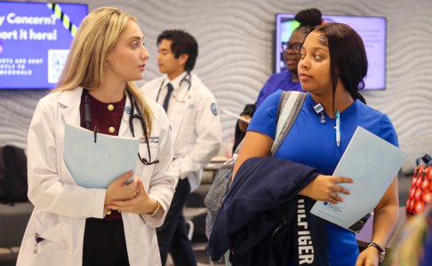 A medical student in a white coat and a nursing student in blue scrubs walk side by side while holding folders. They are engaged in conversation in a busy hallway with other students visible in the background.