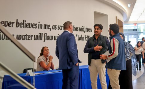 Students and site representatives talk at a BUCOM fair near a wall with the verse John 7:38.