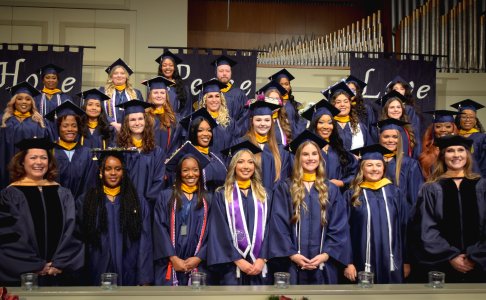 A group of graduates in navy caps and gowns smile on stage during the commencement ceremony, with faculty standing at each end and banners reading “Hope,” “Peace,” and “Love” in the background.