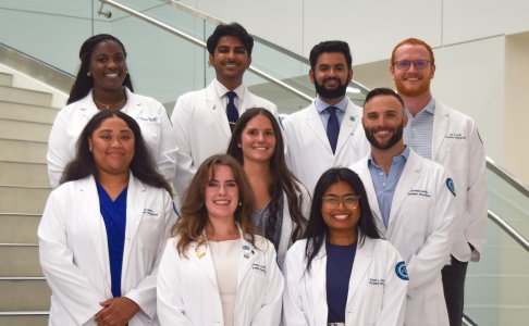 A group of nine BUCOM medical students wearing white coats stand together on a staircase inside a bright, modern building. The students are smiling and posed in two rows, representing the BUCOM Student Government Association.
