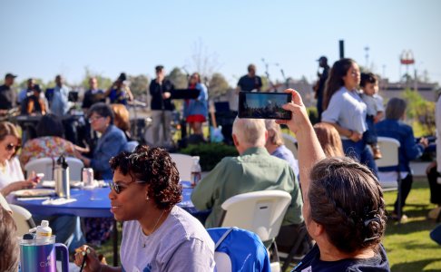 A lively outdoor event with people sitting at tables enjoying food and conversation while a live band performs in the background. A woman in the foreground is taking a photo or video of the band with her phone, and another woman with a name tag reading "Amber" is smiling and chatting. The setting is sunny with clear skies.