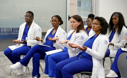 Students dressed in blue scrubs and white coats hold their hands up and out
