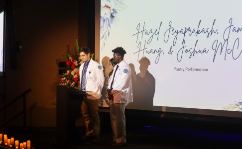 Two medical students wearing white coats stand at a podium on stage during a ceremony. They are presenting in front of a large screen displaying floral graphics and scripted text that reads “Hazel Jeyaprakash, Jam Huang, & Joshua McC… Poetry Performance.” A floral arrangement is visible beside the podium, and candles line the stage steps, creating a formal and reflective atmosphere.