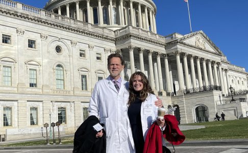 Catie Lockhart in a white coat and a red coat in hand standing with Dr. Bell in a white coat and black coat in hand standing in front of the Capitol Hill building