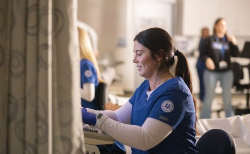 Female sonography student in blue scrubs smiling while using an ultrasound machine in a clinical lab setting, with other students and instructors visible in the background.
