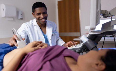 A smiling healthcare professional performs an ultrasound on a patient lying on an exam table. The provider wears a white lab coat and looks at a monitor while holding the ultrasound probe, engaging warmly with the patient.