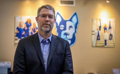 A man with short gray hair, a beard, and glasses wearing a dark blazer and blue shirt stands indoors. Behind him are two blurred wall photos of people in medical scrubs and a large blue husky dog logo.